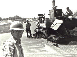 B/w photo of man in hard hat, with construction crew in background